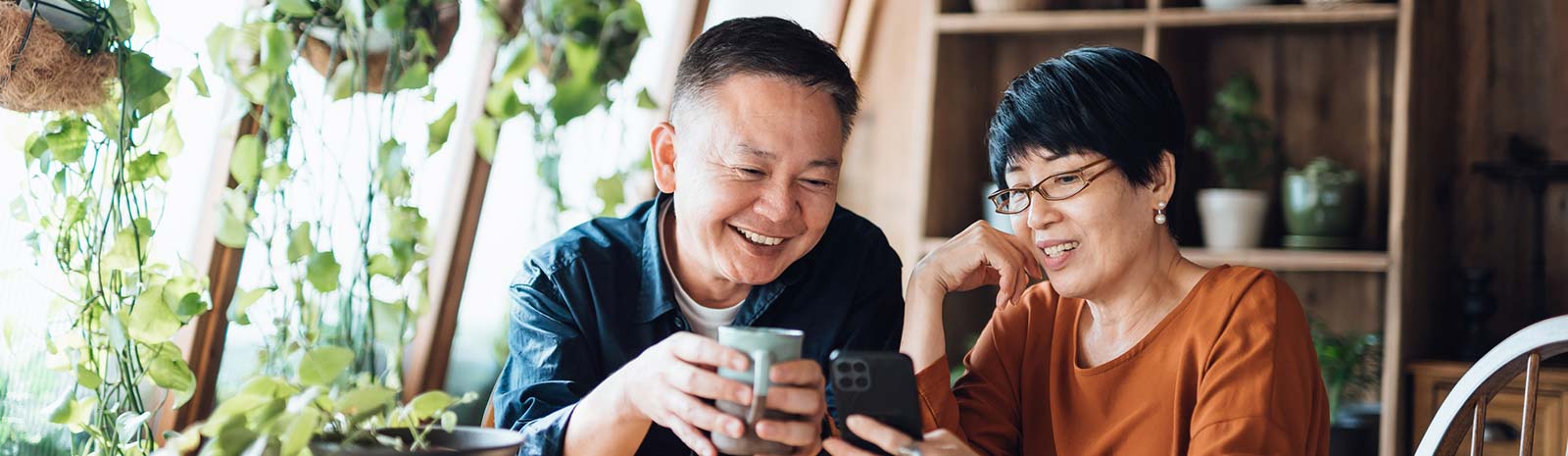 Couple looking at phone and holding coffee in plant-filled room.